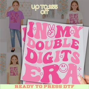 May include: A sheet of pink and light pink "IN MY DOUBLE DIGITS ERA" DTF transfers. The design includes a peace sign, stars, and a smiley face. The image also shows a purple t-shirt and a white t-shirt with the same design.