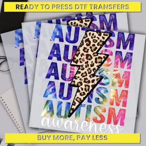 May include: A stack of ready-to-press DTF transfers featuring a rainbow tie-dye background with the word "AUTISM" repeated multiple times. A leopard print lightning bolt graphic is centered on the design. The text "awareness" is printed below the graphic. The text "BUY MORE, PAY LESS" is printed at the bottom of the design.