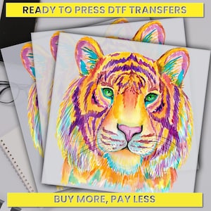 May include: A colorful watercolor illustration of a tiger's face. The tiger has bright green eyes, a pink nose, and purple, orange, and yellow stripes. The background is white. "Ready to Press DTF Transfers" and "Buy More, Pay Less" are written in yellow text at the top and bottom of the image.