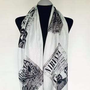 May include: A white scarf with black and grey graphic prints featuring band imagery and text. The scarf is draped over a mannequin, showcasing its design and texture. The text includes "Nirvana" and "MTV's Unplugged".