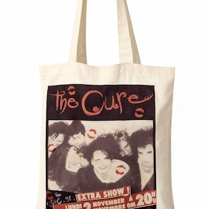 May include: Cream tote bag with a vintage poster design for The Cure's Extra Show. The poster features band members, the band's name, and the show's date and time. The bag has two cream handles. The text includes the words 'Extra Show'.