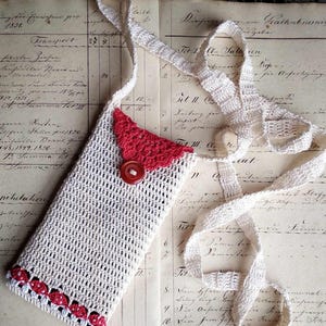 Peut inclure: Une coque de téléphone en crochet avec une bordure rouge et blanche et un bouton rouge. La coque est suspendue à une sangle en crochet blanche.