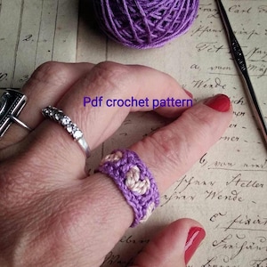 Może przedstawiać: Zbliżenie na dłoń z fioletowym i białym pierścionkiem na szydełku. Pierścień wykonany jest z wzorem w kształcie serca. Na dłoni jest również srebrny pierścień z przezroczystymi kamieniami.