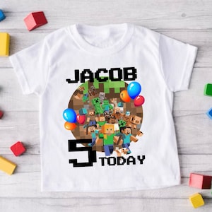 May include: White t-shirt with a Minecraft themed birthday design. The design features a large number "5" with the word "TODAY" below it.  The number is surrounded by Minecraft characters and a green block. The name "JACOB" is printed above the design.