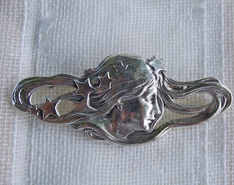 May include: A silver brooch with a stylized image of a woman's face surrounded by stars. The woman's hair flows out behind her head.