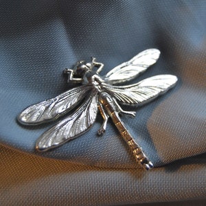 Puede incluir: Broche plateado con forma de libélula, con alas y cuerpo detallados. El broche se encuentra sobre un fondo de tela azul grisáceo suave, resaltando el intrincado diseño y el brillo metálico de la joya. Los delicados detalles de la libélula son claramente visibles.