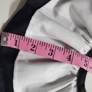 May include: A pink measuring tape with black markings is placed over a white and black fabric. The tape measures 6 inches.