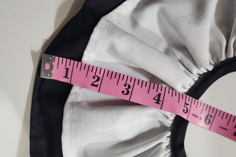May include: A pink measuring tape with black markings is placed over a white and black fabric. The tape measures 6 inches.
