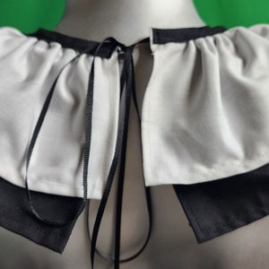 May include: A white ruffled collar with a black ribbon tie. The collar is made of a soft, lightweight fabric and has a simple, elegant design.