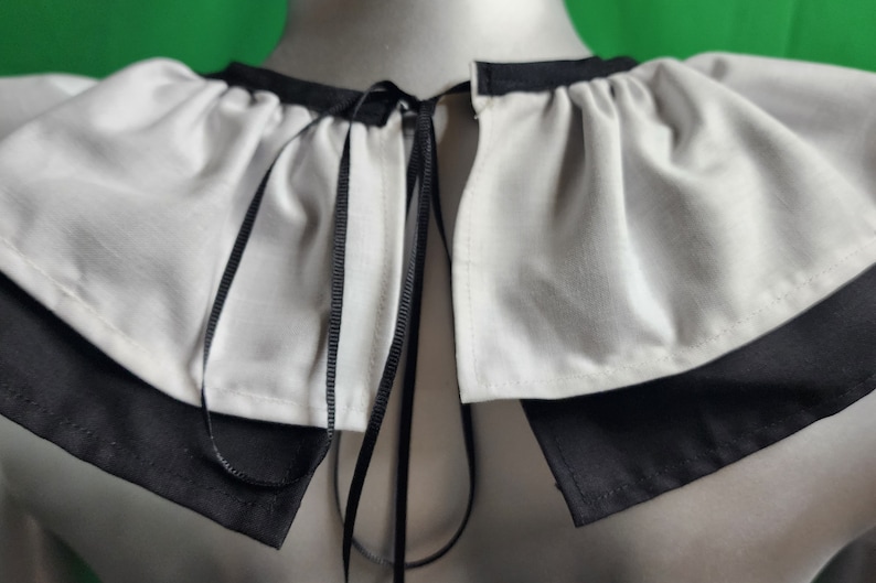 May include: A white ruffled collar with a black ribbon tie. The collar is made of a soft, lightweight fabric and has a simple, elegant design.