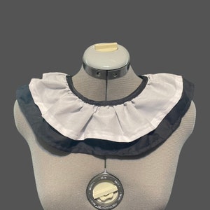 May include: A white ruffled collar with a black trim. The collar is on a dress form.