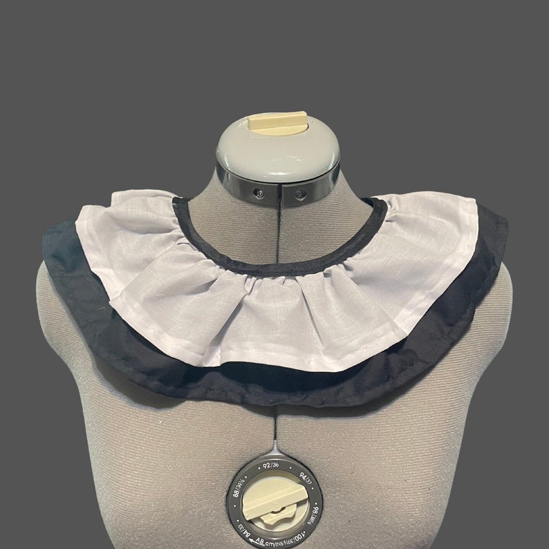 May include: A white ruffled collar with a black trim. The collar is on a dress form.