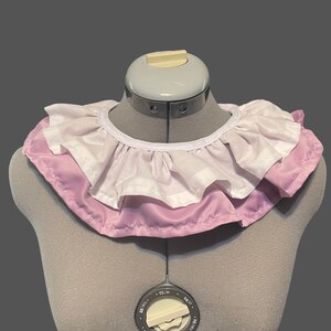 May include: A white ruffled collar with a pink satin trim. The collar is displayed on a dress form.