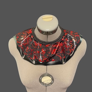 May include: A black and red ruffled collar with a splatter pattern. The collar is made of a shiny fabric and has a black trim.