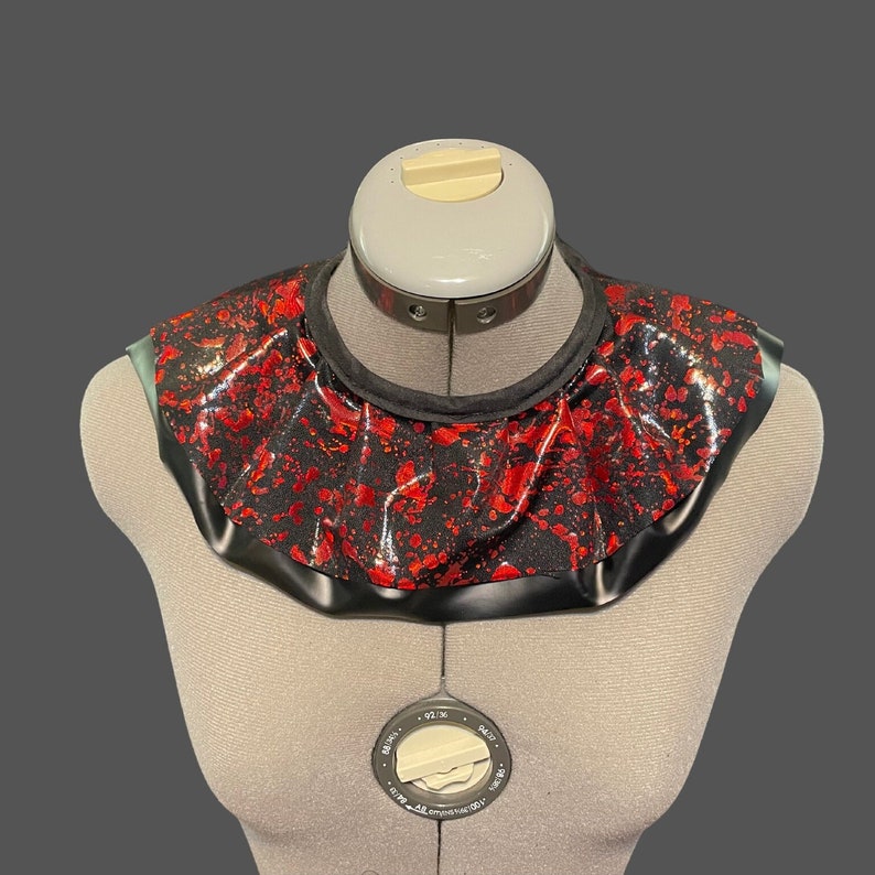 May include: A black and red ruffled collar with a splatter pattern. The collar is made of a shiny fabric and has a black trim.