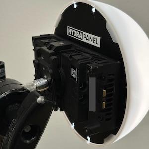 Puede incluir: Un foco Hydra Panel en blanco y negro con un soporte de montaje negro. La luz tiene un difusor blanco y redondeado y está fijada a un soporte negro. El texto "HYDRA PANEL" es visible en el foco.