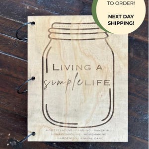 May include: Wooden sign with a mason jar design and the words "Living a Simple Life." The sign includes the phrases "Homesteading, Farming, Ranching, Homeschooling, Homemaking, Gardening, Animal Care." The sign is held together with black metal rings.