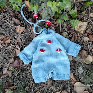 Pode incluir: Um macacão de bebê de tricô azul claro com um design de cogumelo. O macacão tem três cogumelos vermelhos com manchas brancas na frente.