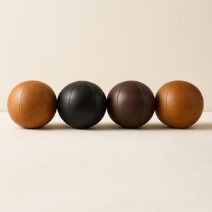 May include: Four leather decorative balls in shades of brown and black. The balls are round with vertical stitched seams. The colors include light brown, black, dark brown, and medium brown. The balls are arranged in a row.