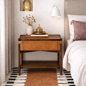 May include: A dark wood bedside table with a drawer featuring a tan leather front. The table has a lower shelf and is next to a bed with a decorative pillow. A vase, books, and a decorative box sit on top.