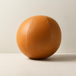 May include: A round, tan-colored leather ball with vertical seams. The ball has a smooth surface and a spherical shape. This decorative ball could be used as a decorative accent piece.