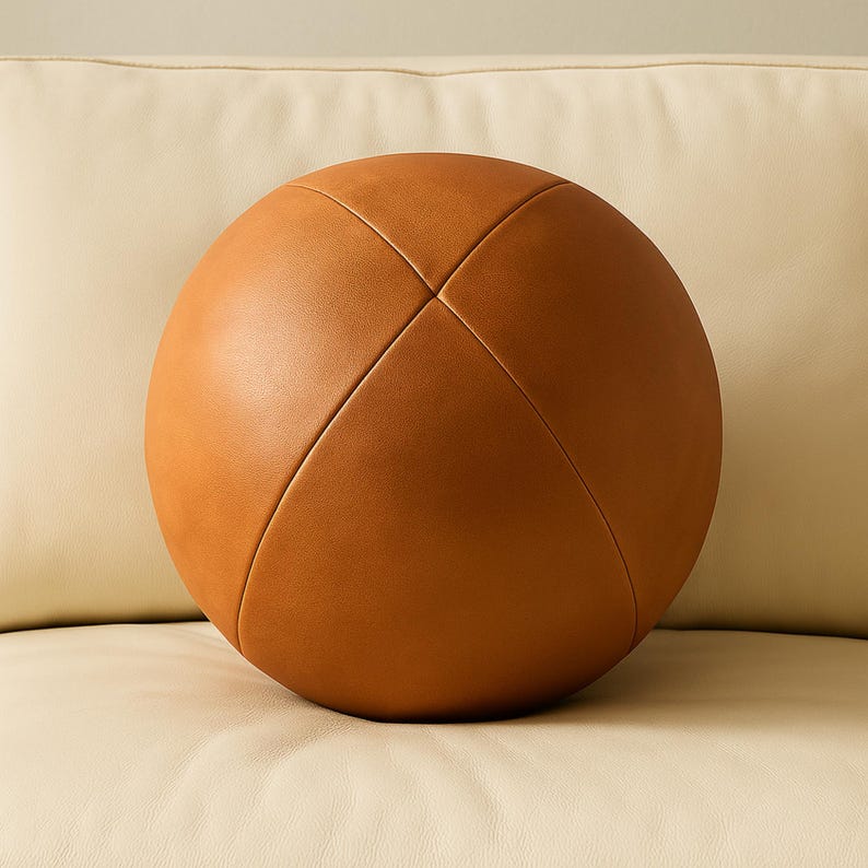 May include: A round, brown leather decorative pillow with a stitched cross pattern. The pillow is a warm, medium brown color and appears to be made of a soft material. The pillow is resting on a cream-colored surface.