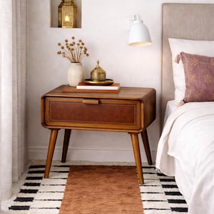 May include: A mid-century modern style nightstand with a brown leather drawer front and wooden frame. The nightstand has tapered legs and a curved top. A small rug with a black and white pattern is in front of the nightstand.
