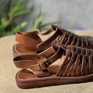 May include: Brown leather sandals with a woven design and a buckle closure. The sandals have a thick, flat sole.