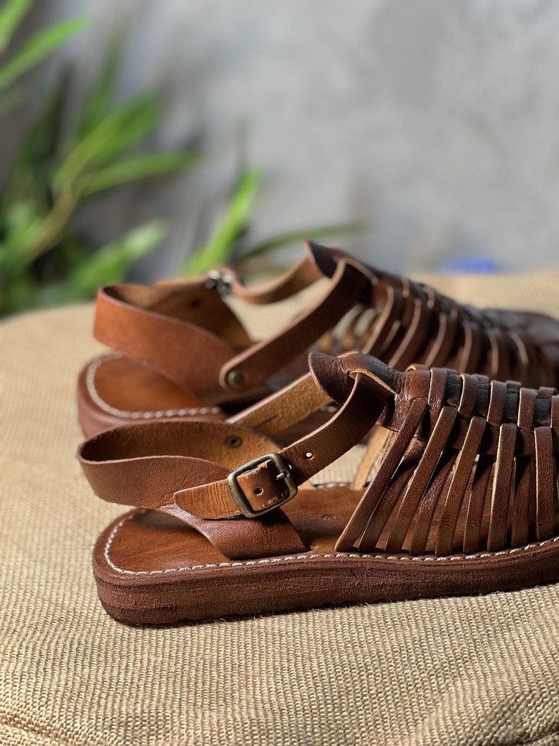 May include: Brown leather sandals with a woven design and a buckle closure. The sandals have a thick, flat sole.