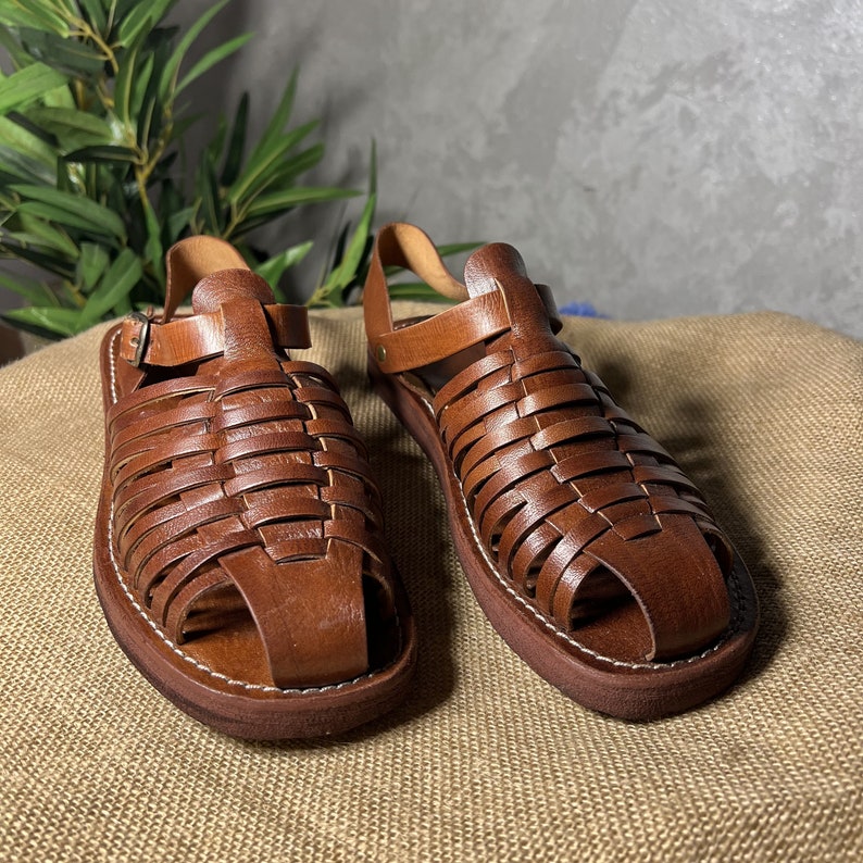 May include: A pair of brown leather sandals with a woven design. The sandals have a strap that goes around the ankle and a buckle closure.