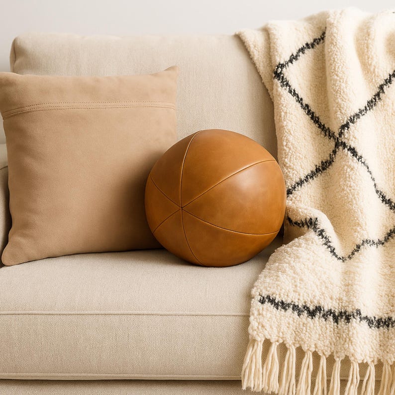 May include: A tan leather decorative ball and a square pillow rest on a beige sofa. A cream-colored throw blanket with a black geometric pattern and fringe is draped over the sofa. The ball has a stitched design.