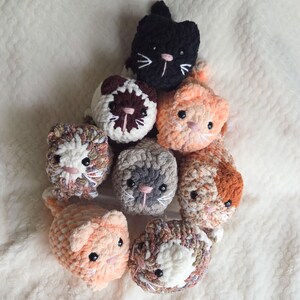 May include: A collection of handmade crochet cat toys in various colors. The cats have embroidered faces with whiskers and small ears. Colors include black, orange, gray, brown, and speckled yarn. The toys are arranged on a soft, white, textured surface.