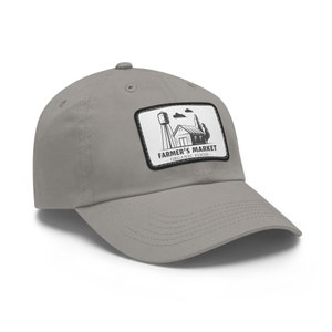 Peut inclure: Casquette de baseball grise avec une visière incurvée. Elle présente un écusson rectangulaire avec une illustration en noir et blanc d'une ferme et le texte "FARMER'S MARKET - ORGANIC FOOD."