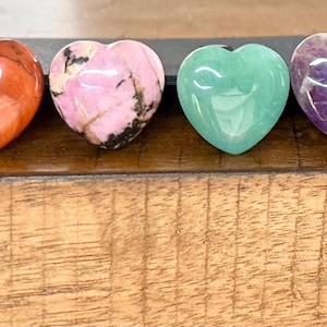 May include: Four heart-shaped stones in shades of red, pink, green, and purple. The stones are polished and smooth, with visible patterns and variations in color. They are arranged on a wooden surface.