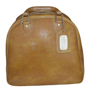 May include: A vintage brown leather travel bag with a rounded top and a handle. The bag has a luggage tag attached with a metal chain. The leather shows signs of wear and tear, adding to its vintage appeal.