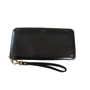 May include: Black leather Fossil wallet with a wrist strap. The wallet has a zipper closure and a gold-colored clasp. The Fossil logo is visible on the top of the wallet.