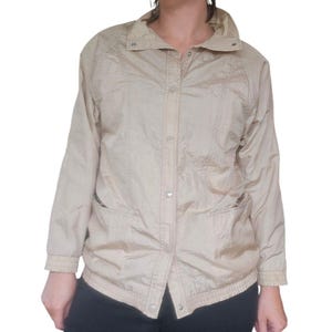 May include: A tan-colored jacket with a collared neckline, button-up front, and two front pockets. The jacket has long sleeves with elastic cuffs and a slightly textured appearance. The overall style is casual and lightweight.