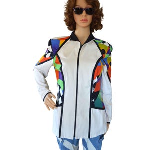 Könnte beinhalten: Weiße Jacke mit Reißverschluss und bunten geometrischen Mustern an Schultern und Seiten. Die Jacke hat einen schwarzen Kragen und Besatz sowie einen vertikalen schwarzen Streifen auf der Vorderseite. Die Ärmel sind lang und weiß.