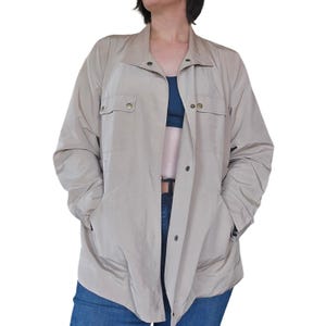 May include: A tan jacket with a button-up front, two chest pockets, and two side pockets. The jacket has a collar and long sleeves. The jacket is open, revealing a dark blue top and blue jeans.