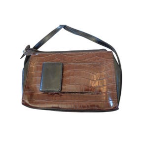 May include: A brown handbag with a crocodile skin texture. The bag has a black handle and a small black flap on the front. The bag has a zipper closure.
