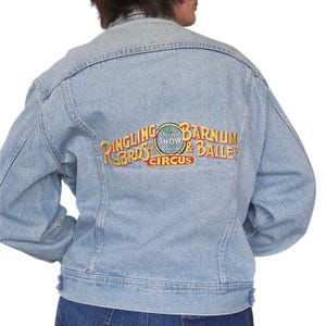 Puede incluir: Chaqueta vaquera azul claro con el logotipo bordado de Ringling Bros. Barnum & Bailey Circus en la espalda. El logotipo incluye las palabras "Ringling Bros" en dorado, "Greatest Show on Earth" en verde y "Barnum & Bailey Circus" en dorado.