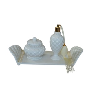 May include: A white vanity set featuring a rectangular tray with flared sides, a lidded jar, and a perfume bottle with a gold pump and cream-colored tassel. Each piece has a quilted texture with small pearl-like accents along the edges.