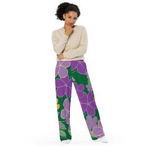May include: Loose-fitting pants with a vibrant floral pattern. The pants feature large purple flowers with yellow centers and green leaves against a dark green background. The model is wearing a light beige long-sleeved shirt.