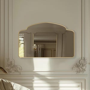 Könnte beinhalten: Ein elegantes Interieur mit einem goldgerahmten Spiegel, einem cremefarbenen Sessel und einem Kamin. Die Wände weisen detaillierte Formteile und dekorative Elemente auf. Eine Vase mit getrockneten Pflanzen befindet sich in der Nähe des Stuhls, und ein Teppich liegt auf dem Boden.