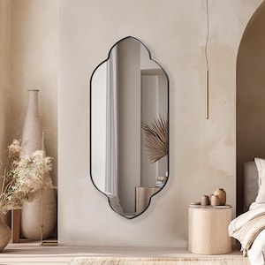 May include: A neutral-toned bedroom with a large arched doorway and a bed covered in white and beige bedding. A black-framed mirror hangs on the wall. A beige rug and decorative vases complete the space.