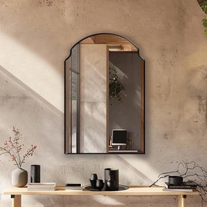 以下が含まれることがあります： シンプルな黒いフレームのアーチ型の黒い鏡が、薄いベージュの壁に、木製のコンソールテーブルの上にかけられています。テーブルには、赤い実の入った花瓶、黒い花瓶、本の束が置かれています。