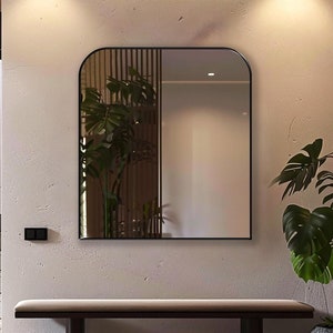 Pode incluir: Um espelho arqueado com moldura preta pendurado em uma parede bege claro acima de um banco de madeira. Uma planta em vaso com grandes folhas verdes fica no chão ao lado do banco. Três luzes de globo preto penduradas no teto.