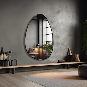 May include: A black oval mirror with a thin black frame hangs on a gray concrete wall. A wooden bench with black legs sits in front of the mirror. A black round ottoman sits on a gray rug in front of the bench. A potted plant is in the background.