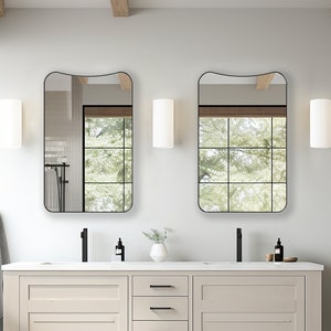 Puede incluir: Un tocador de baño blanco con dos lavabos y dos grifos negros. El tocador tiene tres cajones con tiradores negros. Dos espejos negros enmarcados con bordes ondulados cuelgan sobre los lavabos. Los espejos reflejan una vista de los árboles fuera de la ventana.