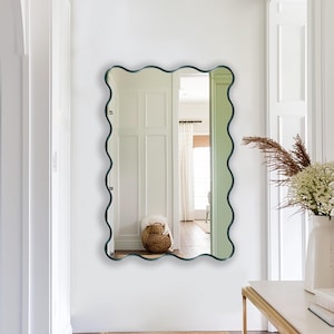 May include: A green wavy framed mirror hangs on a white wall in a hallway with a gold console table and a vase of dried flowers.